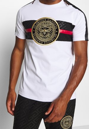 Homme portant un t-shirt blanc avec une bande noire et rouge et un emblème d'abeille doré « Glorious Gangsta », associé à un short noir à motif avec un emblème assorti.