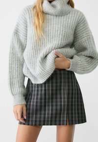 Femme portant un pull à col roulé épais gris clair et une mini-jupe à carreaux noire et grise avec une petite fente à l'avant.