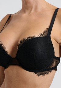 Sostenedor de encaje negro con un patrón floral, copas acolchadas y tirantes ajustables, con un borde texturizado y con forma de concha a lo largo de los bordes.