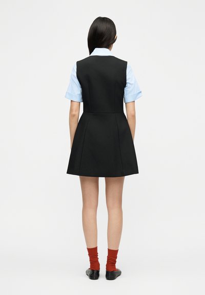 Γυναίκα με μακριά σκουρόχρωμα μαλλιά, ντυμένη με ανοιχτό μπλε μπλουζάκι κοντομάνικο κάτω από μαύρο φόρεμα χωρίς μανίκια, κόκκινες κάλτσες και μαύρα παπούτσια, στέκεται με την πλάτη προς τα εμάς.