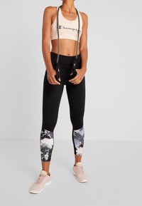 Kobieta w czarnych legginsach w kwiaty, jasnoróżowym staniku sportowym i trampkach trzymająca czarny skakankę w tle na jednolitym tle.