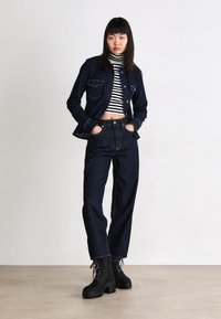 Ciemna kurtka dżinsowa z dwoma przednimi kieszeniami, w połączeniu z ciemnymi dżinsami z wysokim stanem. Pod spodem biało-czarny golf w paski. Masywne czarne buty.