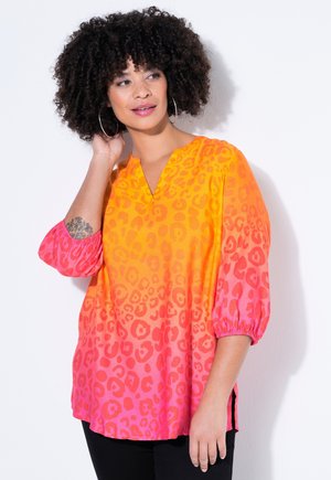 Kvinde med krøllet hår iført øreringe i ringform og en langærmet bluse med leopardprint i orange til pink gradient, der rører ved sit hår med den ene hånd.