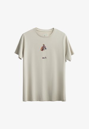 T-shirt de couleur crème en coton doux, avec un motif graphique central représentant un personnage portant une cape marron et des accents bleus.