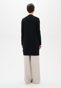 Lang zwarte cardigan van zacht materiaal, met lange mouwen en een rechte snit, gecombineerd met lichtbeige wijdvallende broek.