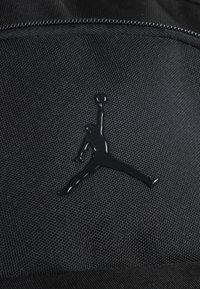 Schwarzer Stoffrucksack mit einem glänzenden schwarzen Jumpman-Logo, einer strukturierten Webmusterung und kontrastierenden glatten Reißverschlussdetails.