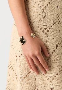 Pulsera en tono dorado con un diseño escultórico abstracto y superficies pulidas; lleva en una mano sobre un tejido de ganchillo de color crema.