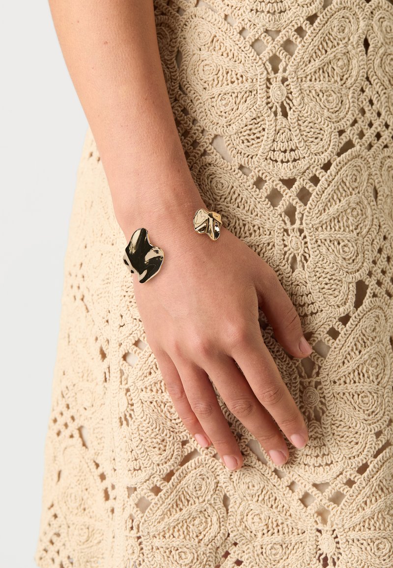 Pulsera en tono dorado con un diseño escultórico abstracto y superficies pulidas; lleva en una mano sobre un tejido de ganchillo de color crema.