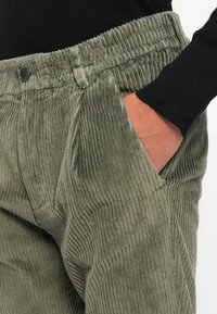 Ruka odmara u džepu zelenih kordura hlača s elastičnom trakom u struku, nosi crni rebrasti majicu dugih rukava.