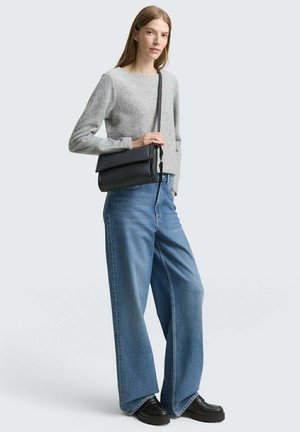 Grauer Wollpullover, weite blaue Jeanshose und schwarze Umhängetasche. Das Model trägt klobige schwarze Schuhe; der neutrale Hintergrund hebt die Details der Kleidung hervor.