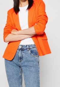 Blazer ajusté orange vif à manches retroussées, associé à un haut blanc côtelé et un jean taille haute bleu clair avec fermeture par boutons.
