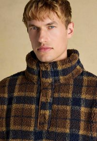 Brown and blue plaid fleece pullover with a high collar, featuring a buttoned placket and textured fabric.