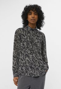Zwarte en crèmekleurige blouse met lange mouwen en een hoge kraag, met een golvend patroon. Lichtgewicht stof met een relaxte pasvorm, gestopt in grijze broeken.