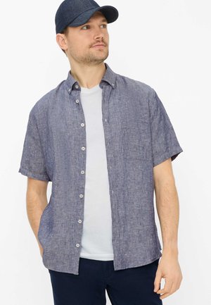 Mand iført en blå kortærmet skjorte med knapper over en hvid t-shirt og en marineblå baseballkasket, der kigger lidt til siden.