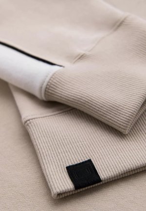 Sudadera beige con puños acanalados, que presenta una capa interior blanca plana y una pequeña etiqueta negra, mostrando una textura suave y un diseño sencillo.