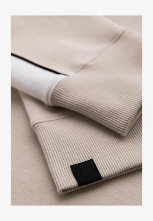 Beige Sweatshirt mit gerippten Bündchen, featuring einer flachen weißen Innenschicht und einem kleinen schwarzen Etikett, zeigt eine weiche Textur und ein schlichtes Design.