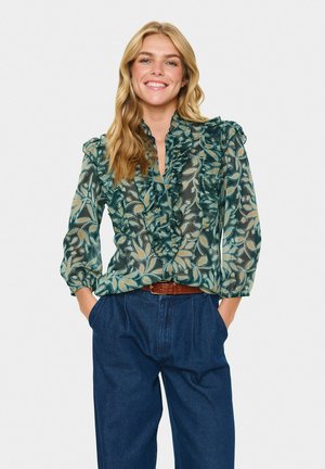 Frau mit langen, welligen blonden Haaren, die lächelt und eine grüne, florale Rüschbluse mit Dreiviertelärmeln und blauen, weit geschnittenen Hosen trägt.