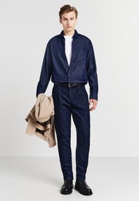 Camicia in denim blu scuro, t-shirt bianca, pantaloni blu scuro, cintura nera, scarpe nere; giacca beige appoggiata sul braccio. Design semplice e su misura.