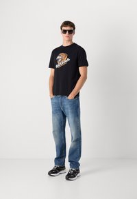 Tricou negru cu mânecă scurtă, având un grafic cu leu, asortat cu blugi albaștri deschiși. Modelul poartă ochelari de soare negri și adidași negri.