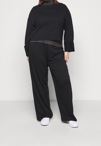 Top corto negro de manga larga superpuesto sobre una camiseta gris oscura, combinado con pantalones negros holgados. Tejido cómodo; diseño sencillo. Zapatos blancos.