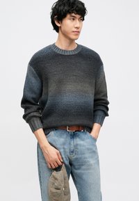 Temno sivi pleteni pulover z gradientnim vzorcem, okroglim izrezom in rebrastimi manšetami. Nošen preko svetlo modrih kavbojk z rjavim pasom.