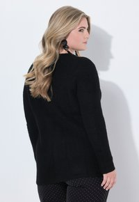 Blonďatá žena s vlnitými vlasy, oblečená v černém svetru, černých kalhotách s puntíky a velkých černých náušnicích, stojící bokem proti bílé zdi.