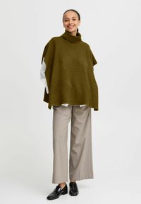 Poncho lavorato a maglia verde oliva con colletto alto, abbinato a pantaloni beige a righe e a gamba larga e mocassini neri, caratterizzato da una vestibilità comoda.