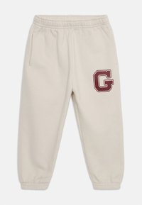 Lys beige sweatpants med elastisk talje, sidelommer, og en maroon bogstav "G" patch på venstre lår. Blød stoftekstur.