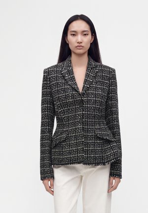 Femme portant un blazer ajusté à carreaux noir et blanc avec deux boutons, associé à un pantalon blanc, debout devant un fond uni.