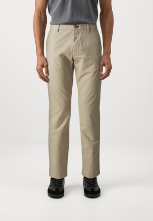 Beige Chinos mit geradem Schnitt, ausgestattet mit einem Knopfverschluss, Seiten Taschen und getragen mit schwarzen Sneakers. Glattes, strapazierfähiges Material.