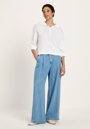 Frau steht vor einfachem Hintergrund, trägt weite hellblaue Jeans, weißes Polohemd mit dreiviertellangen Ärmeln und weiße flache Schuhe.