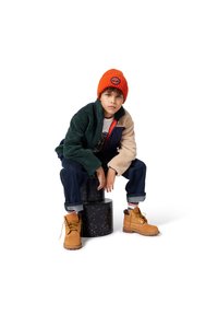 Groene en beige fleece jas met oranje accenten, wit T-shirt, donkere jeans, oranje gebreide muts en bruine veterschoenen.