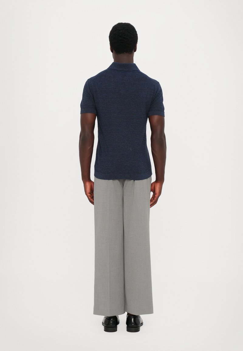 Polo navy a maniche corte con tessuto texturizzato, abbinato a pantaloni larghi grigi chiari e calzature nere. Vista posteriore.
