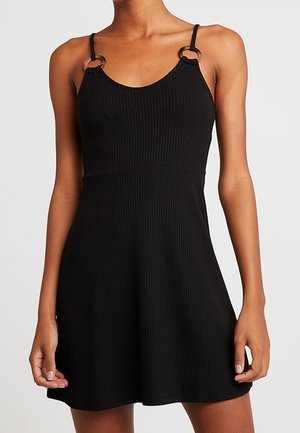 Zwarte geribde mouwloze jurk met dunne bandjes die vastzitten met ronde ringdetails, gedragen door een persoon die tegen een effen achtergrond staat.