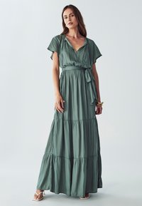 Zelené maxi šaty vyrobené z lehké tkaniny, s krátkým rukávem, V-výstřihem, páskem v oblasti pasu a vrstveným lemem. Hladká textura s jemným leskem.