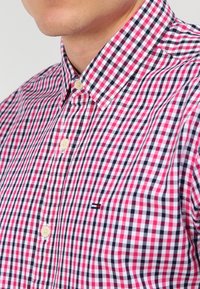 Camicia a quadri rossi e blu con bottoni, realizzata in tessuto dalla texture liscia. Presenta un colletto classico e un piccolo dettaglio del logo sul petto.