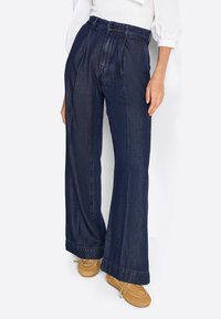 Pantalon en denim bleu foncé, avec une taille haute, un design plissé, des jambes larges et des ourlets retroussés. Associé à des mocassins marron clair.