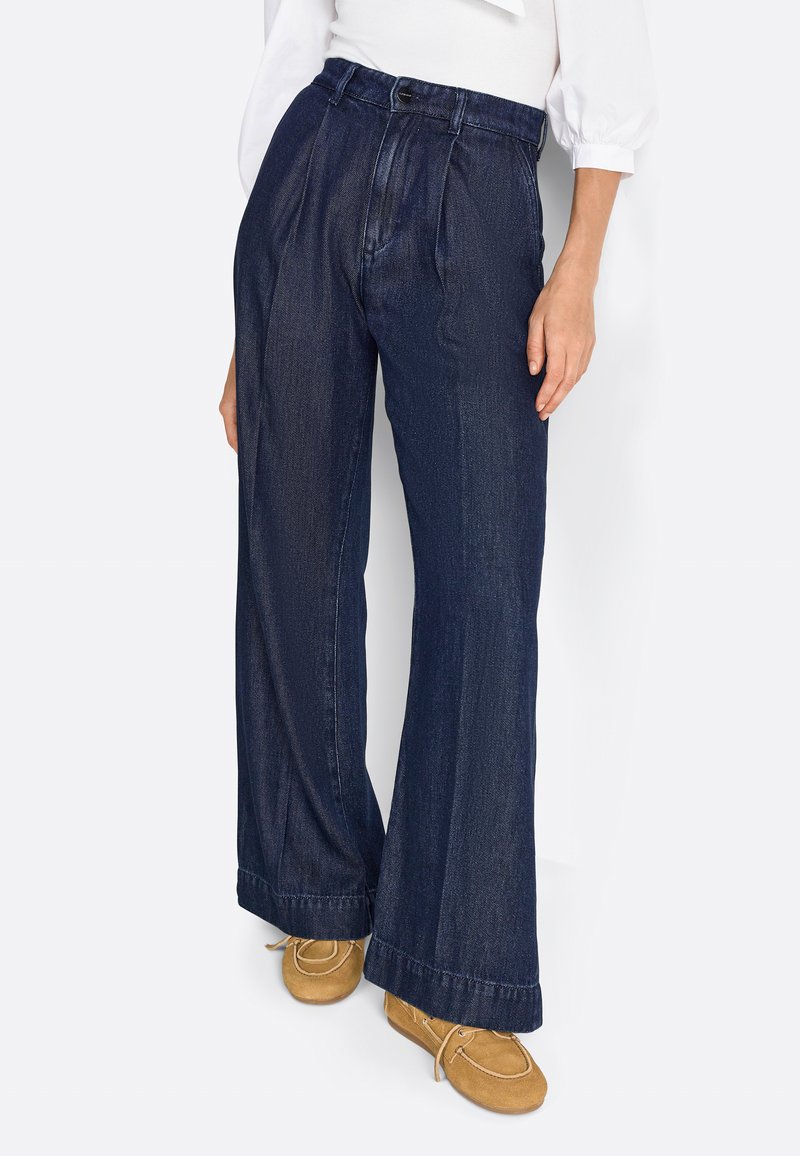 Pantalon en denim bleu foncé, avec une taille haute, un design plissé, des jambes larges et des ourlets retroussés. Associé à des mocassins marron clair.