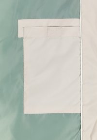 Patch en tissu rectangulaire beige cousu sur un tissu vert avec une double couture et un petit onglet rectangulaire cousu sur le dessus.