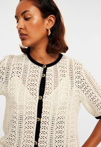 Sweter z dzianiny w kolorze kremowym z teksturowanym wzorem, z krótkimi rękawami, czarnym wykończeniem i złotymi guzikami z przodu.