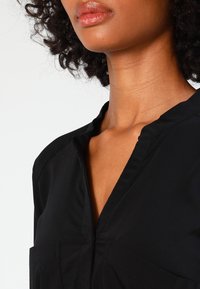 Schwarze Bluse mit V-Ausschnitt, kurzen Ärmeln und zwei Fronttaschen. Der Stoff wirkt glatt und leicht.