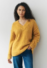 Gul, chunky strikket sweater med V-halssnit og ribbede manchetter, parret med blå jeans. Sweateren har en tekstureret, oversized pasform.