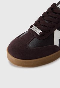 Σκούρο καφέ σουέντ και δερμάτινο αθλητικό παπούτσι με λαστιχένια σόλα, αντίθετες λευκές λεπτομέρειες και ασημένια ετικέτα με λογότυπο στη γλώσσα.