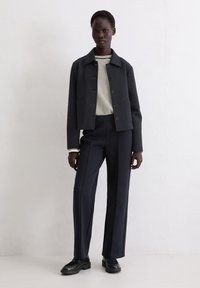 Granatowa marynarka z kołnierzem i guzikami z przodu, zestawiona z pasiastą koszulką z okrągłym dekoltem oraz dopasowanymi, eleganckimi spodniami. Czarne buty dopełniają stylizację.