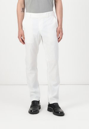 Man draagt witte op maat gemaakte broek, zwarte leren nette schoenen en een grijs ingestopt overhemd, staand tegen een effen witte achtergrond.