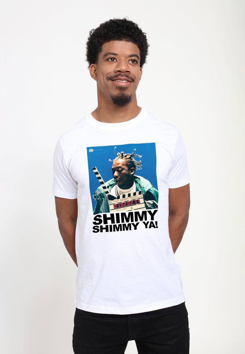 Hvid bomulds t-shirt med et grafisk print af en person med fletninger, iført en grøn jakke og en filmklapper. Teksten siger "SHIMMY SHIMMY YA!"