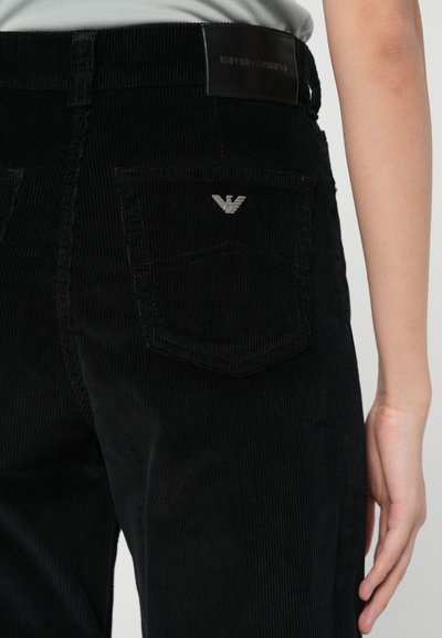 Pantaloni din catifea neagră cu o suprafață texturată, având două buzunare la spate și un mic logo argintiu pe buzunarul din dreapta.