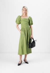 Robe en lin vert avec des manches bouffantes, un corsage froncé et une jupe évasée. Le mannequin tient un sac à main noir texturé, associé à des talons aigus noirs.
