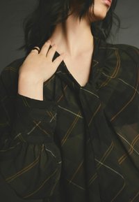 Blouse à carreaux noirs avec des manches bouffantes, présentant un col noué et un tissu doux et transparent. Comprend des lignes d'accent dorées et brunes dans le motif.