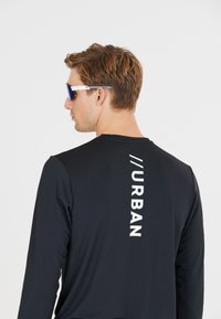 Zwart shirt met lange mouwen van een gestructureerde stof, met een witte verticale "URBAN" print op de achterkant. Gecombineerd met stijlvolle zonnebrillen.